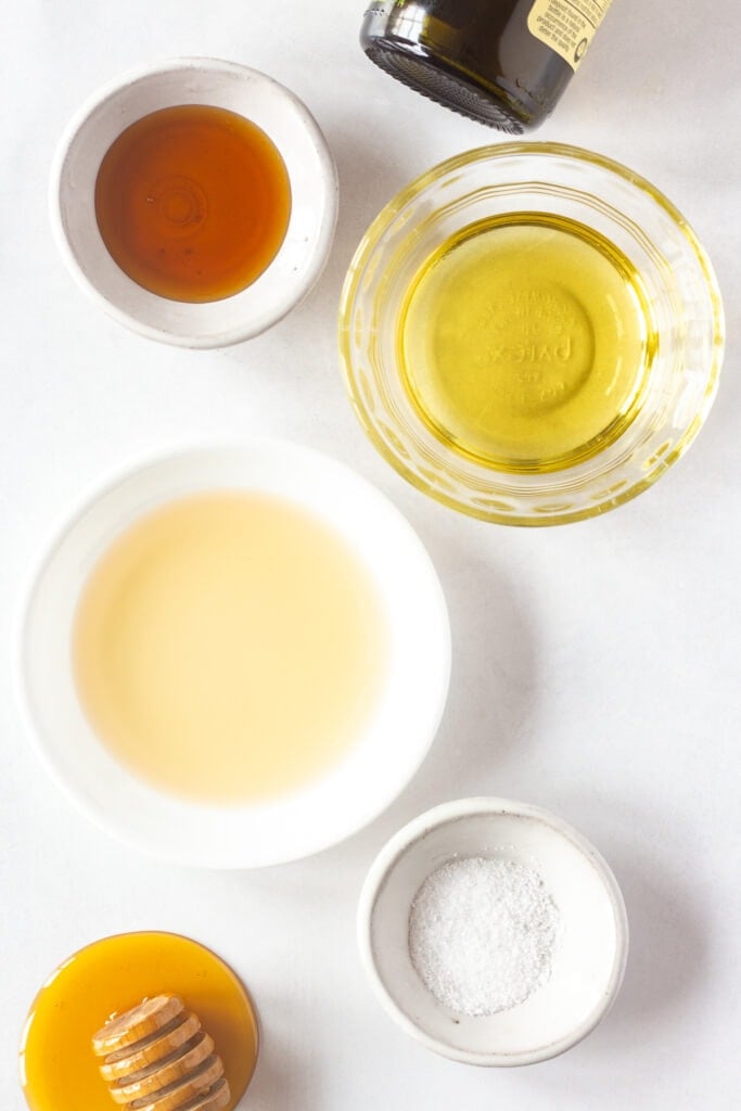 Top down shot of small bowls with honey, oil, vinegar, and kosher salt in them, on a white background.