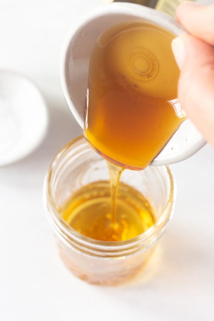 Close up of pouring honey from a small prep bowl into a mason jar with oil and vinegar already in it.