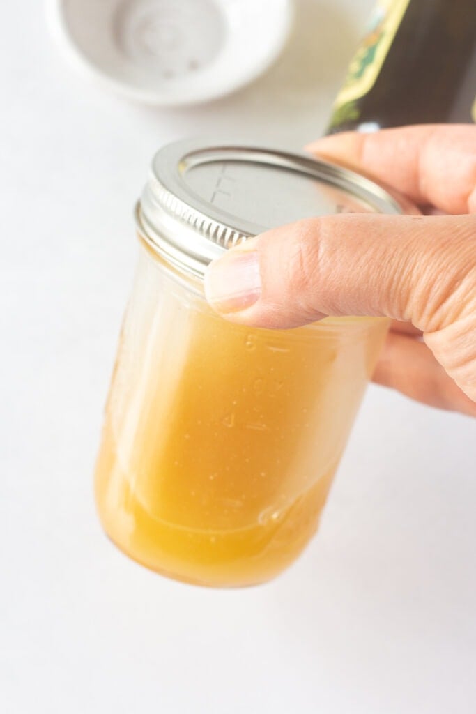 A hand shaking a small lidded mason jar with a gold-ish salad dressing in it.