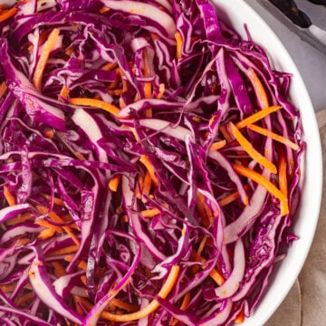 Straight down shot of ¾ of a large white bowl with a purple cabbage and carrot slaw in it. Half an orange is next to it, along with a tan cloth napkin and a pair of tongs.