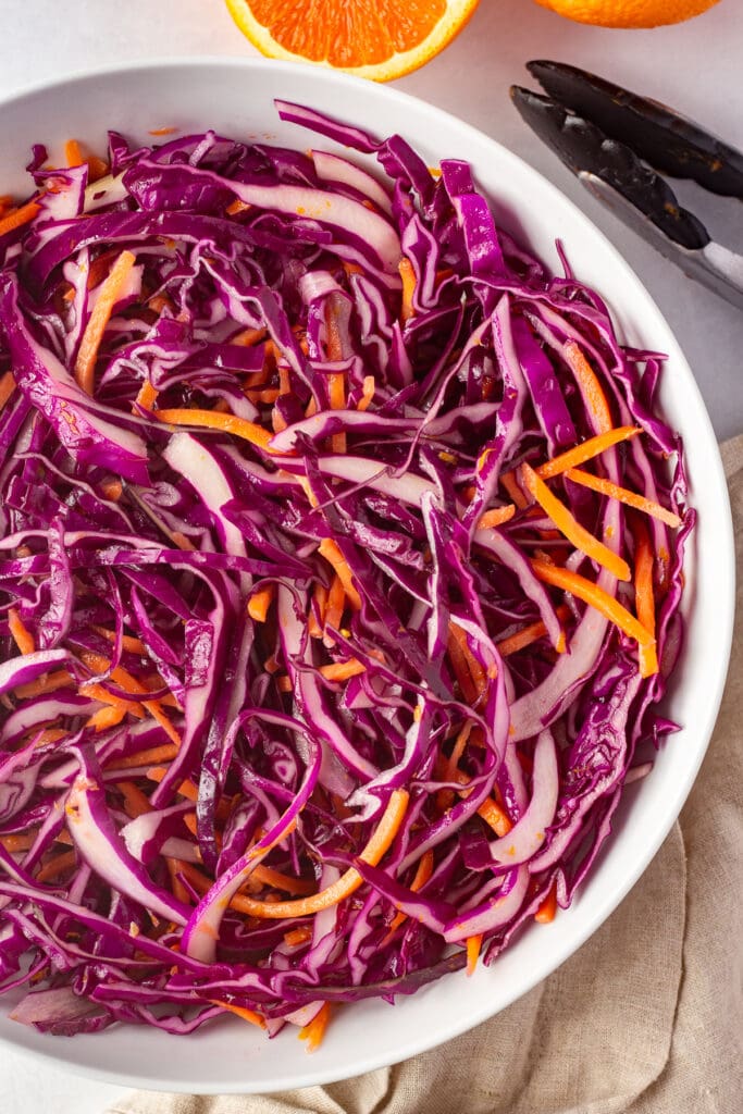 Straight down shot of ¾ of a large white bowl with a purple cabbage and carrot slaw in it. Half an orange is next to it, along with a tan cloth napkin and a pair of tongs.