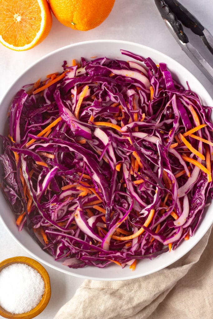 Top down shot of a large white bowl with purple cabbage and carrot slaw in it. A cut orange, a pair of kitchen tongs, a tan cloth napkin, and a small wooden bowl with salt are spread around next to it.