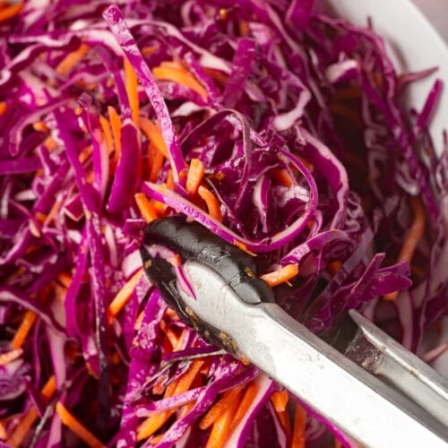 Close up of a pair of kitchen tongs grasping some purple cabbage slaw out of a white bowl.