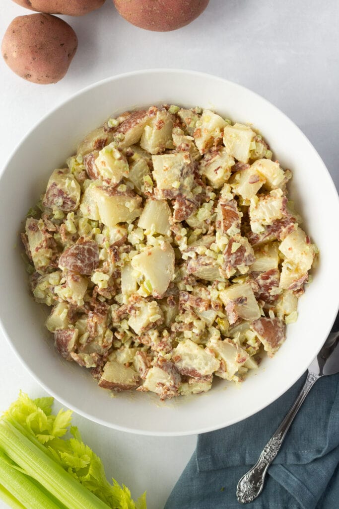Top down shot of bacon red skin potato salad in a large white bowl with a dark blue napkin and serving spoon off the the bottom right of the photo.