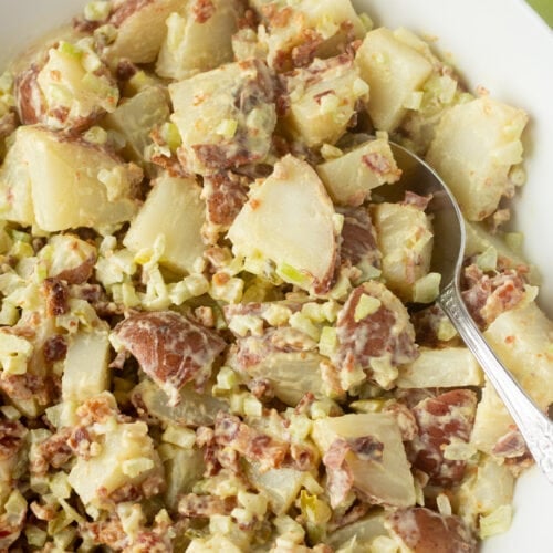 Red skin potato salad with bacon, pickle, and celery in a large white bowl with a serving spoon in it on the right hand side.