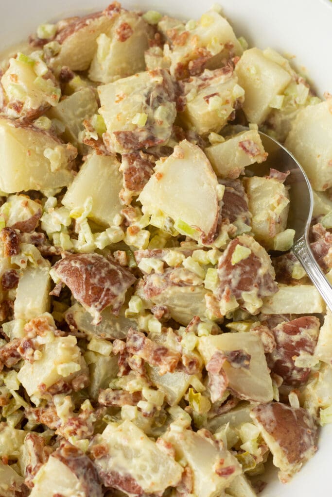 Close up of bacon red skin potato salad in white bowl with a spoon to the right side.
