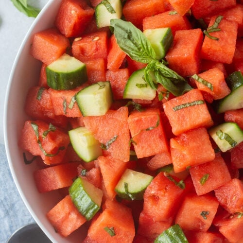 Close up of a bowl of watermelon salad with basil and cucumber in it. The bowl sits on a white background with fresh basil to the top left and a serving spoon in the bottom right of the frame.