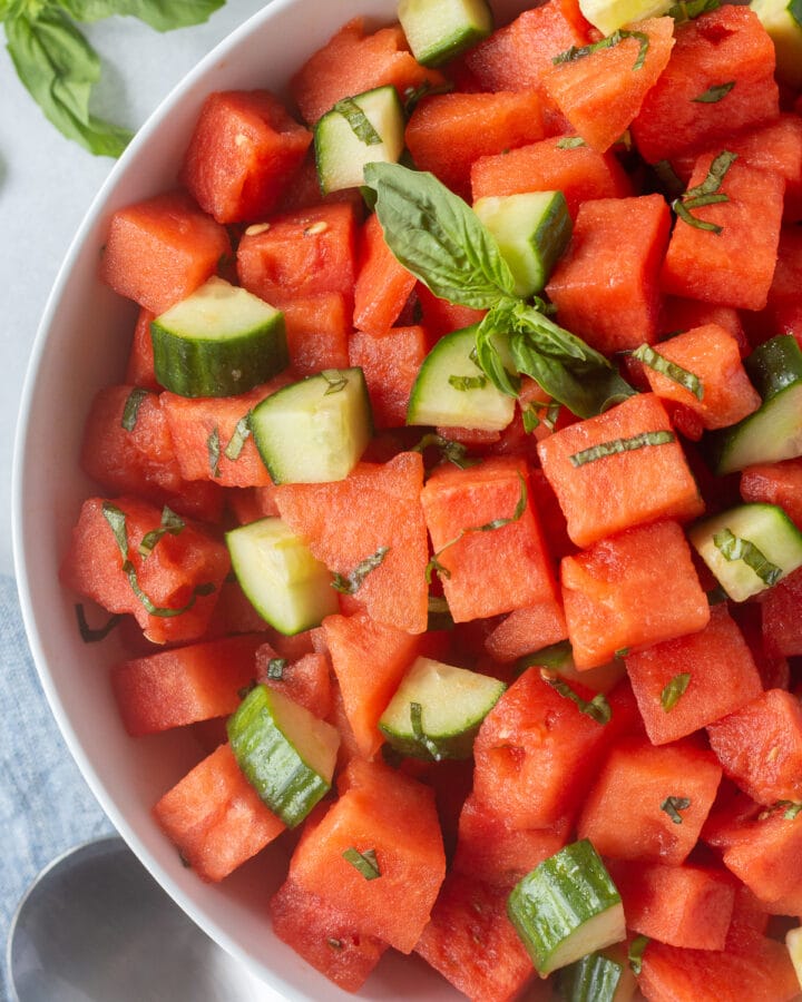 Close up of a bowl of watermelon salad with basil and cucumber in it. The bowl sits on a white background with fresh basil to the top left and a serving spoon in the bottom right of the frame.