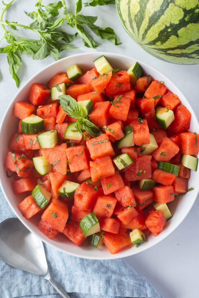 Top down shot of a large white bowl with a watermelon cucumber basil salad in it, surrounded by fresh basil, a watermelon, a blue cloth napkin, and a silver serving spoon.
