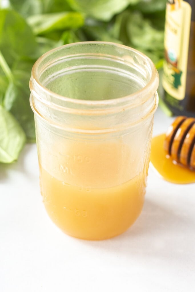 Close up of a small mason jar with a gold-ish dressing surrounded by baby spinach, a vinegar bottle, and a honey stick with honey dripping off it.