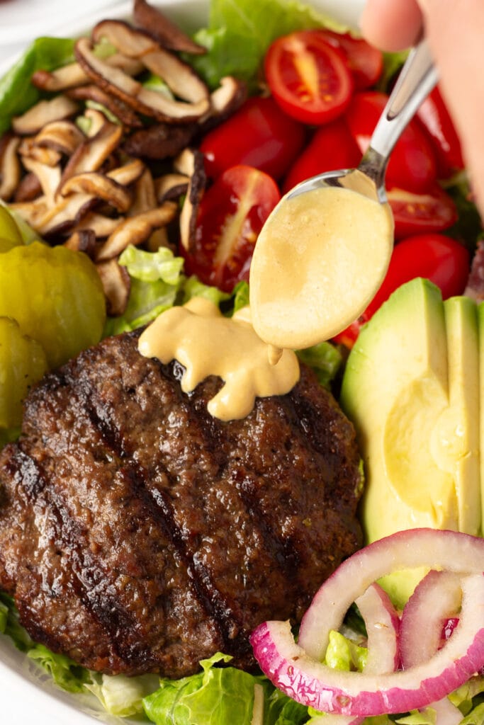 Close up of a golden burger sauce being spooned on top of a burger patty on a salad with mushrooms, tomatoes, avocado, onions, and pickles on it.