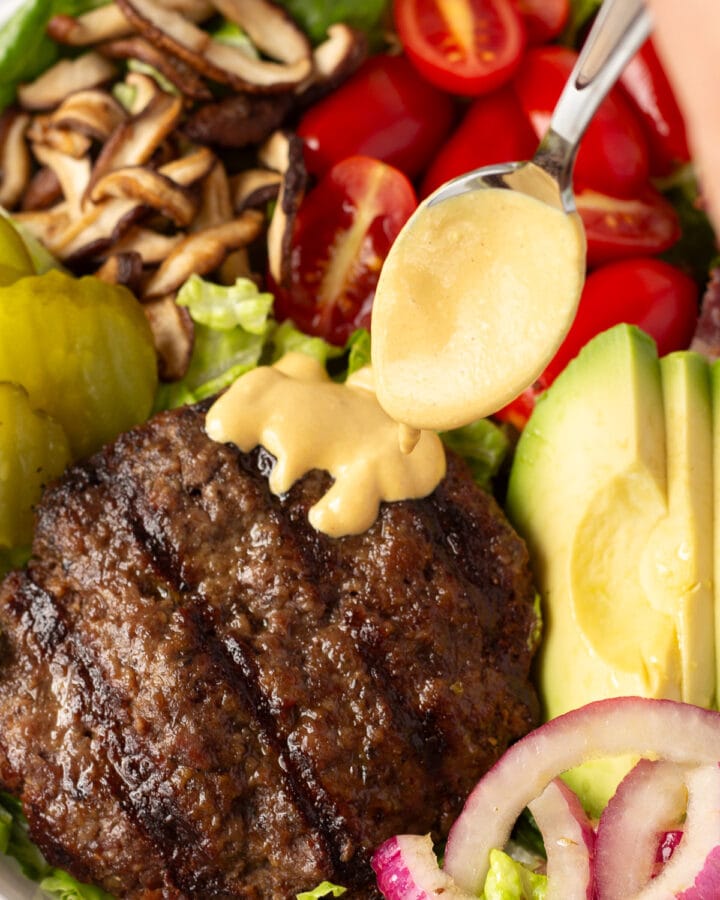 Close up of a golden burger sauce being spooned on top of a burger patty on a salad with mushrooms, tomatoes, avocado, onions, and pickles on it.