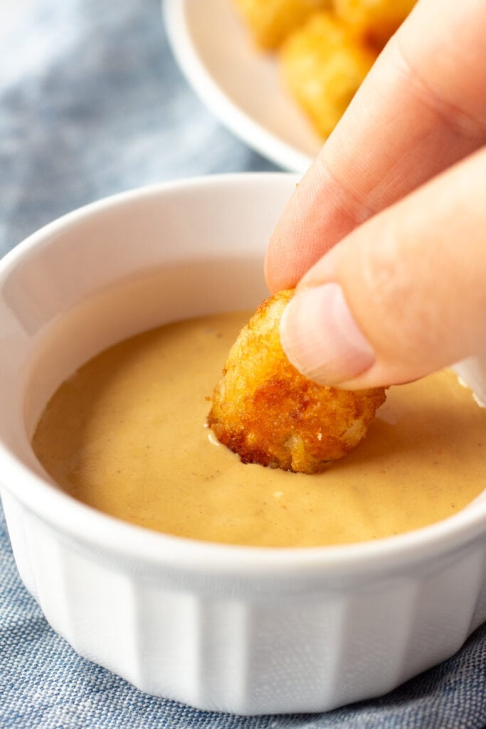 Close up of a hand dipping a tater tot into a golden burger sauce in a small white dish.