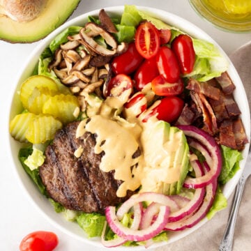 Top down shot of a deconstrcuted burger salad in a large white bowl with ingredients like avocado, tomatoes, pikcles, and bacon off to the side.