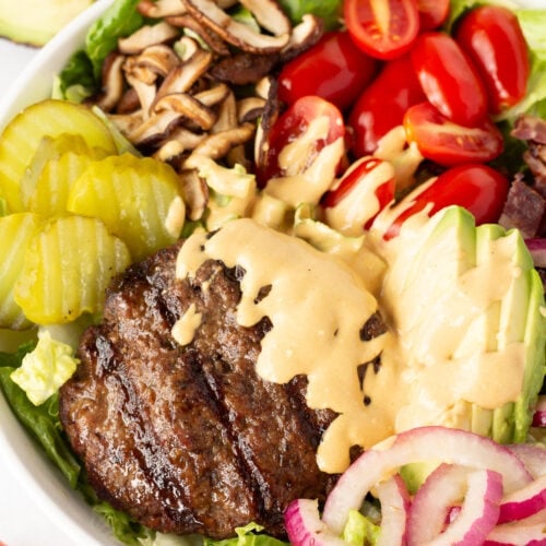 Close up of a deconstructed burger bowl with a beef patty, sliced cherry tomatoes, avocado, red onion, sauteed mushrooms, and pickles in a large white bowl with an orange sauce drizzles over the patty.