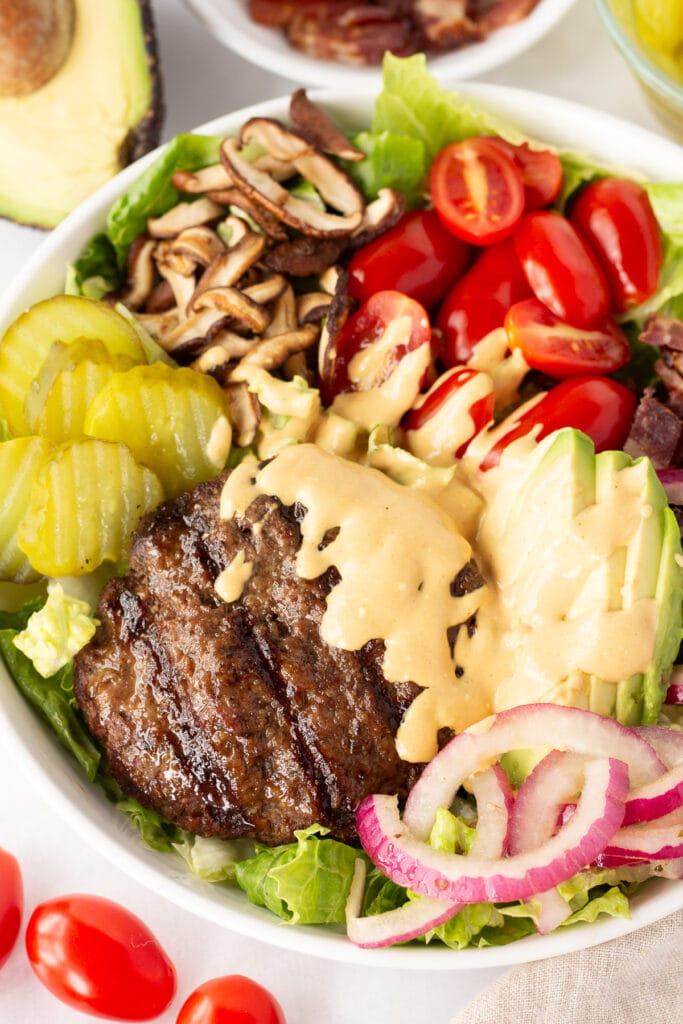 Close up of a deconstructed burger bowl with a beef patty, sliced cherry tomatoes, avocado, red onion, sauteed mushrooms, and pickles in a large white bowl with an orange sauce drizzles over the patty.