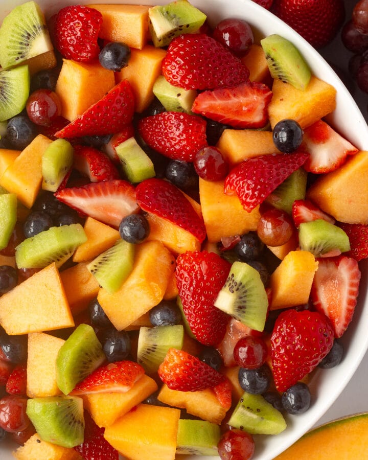 Top down shot of a white bowl with mixed fruit salad in it, surrounded by fresh fruit on the sides.