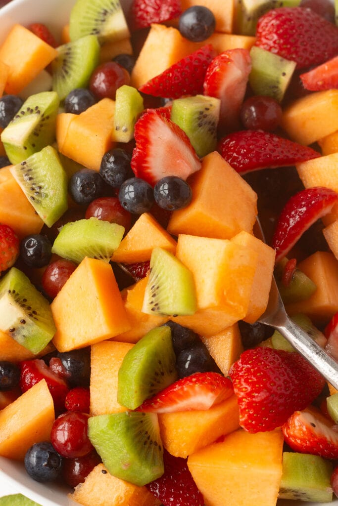 Close up of fruit salad being mixed by a large silver spoon.
