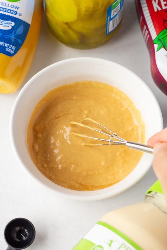 Close up of a hand using a small whisk to stir a golden sauce in a small white dish.