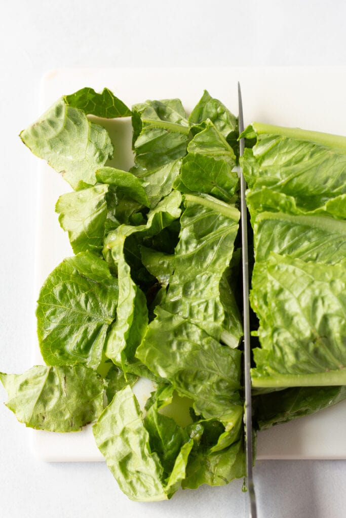 Close up of a large knife cutting a head of romaine lettuce into small pieces on a white cutting board.