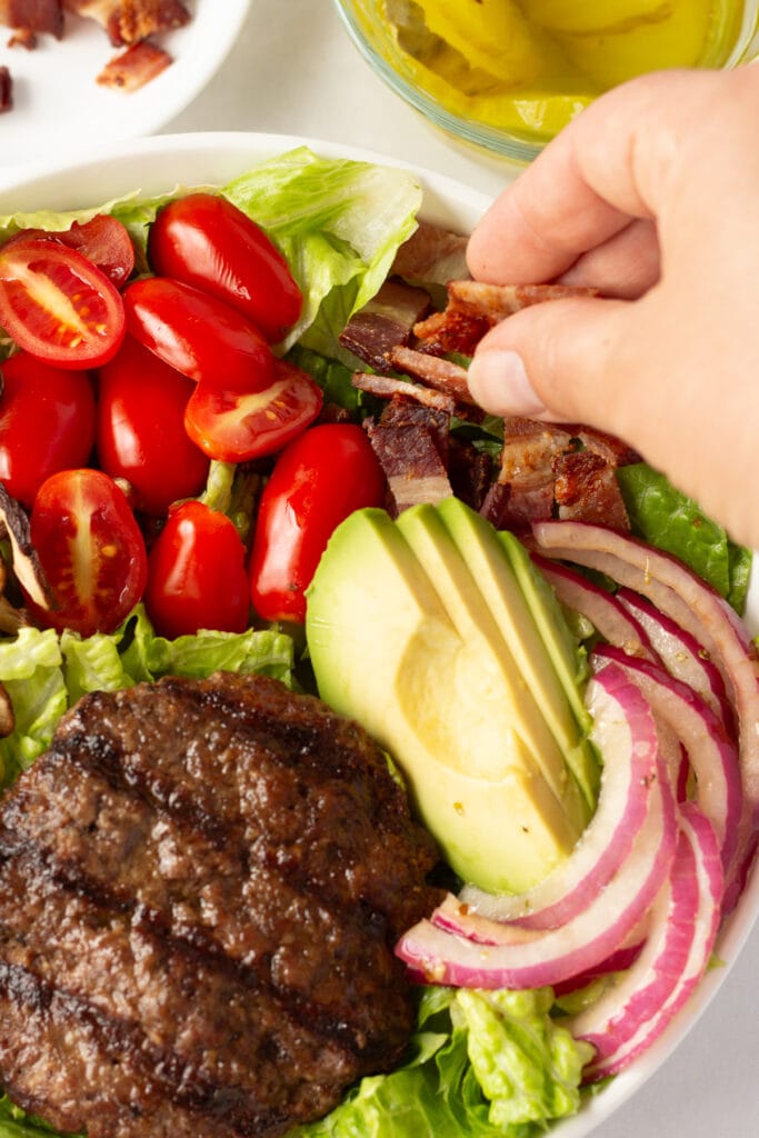 Close up of a hand adding sliced cooked bacon pieces to a bowl with lettuce, avocado, onion, tomatoes, and a grilled burger in it.
