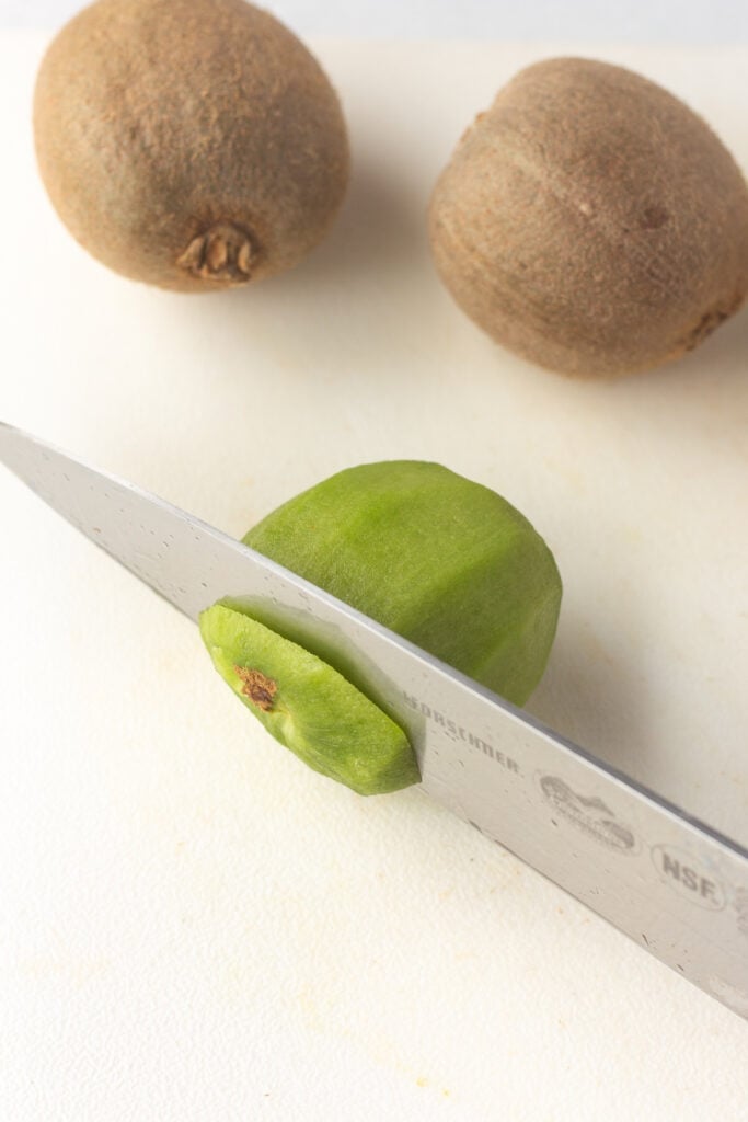 Cutting the skinned end of a kiwi off with a large knife on a cutting board.