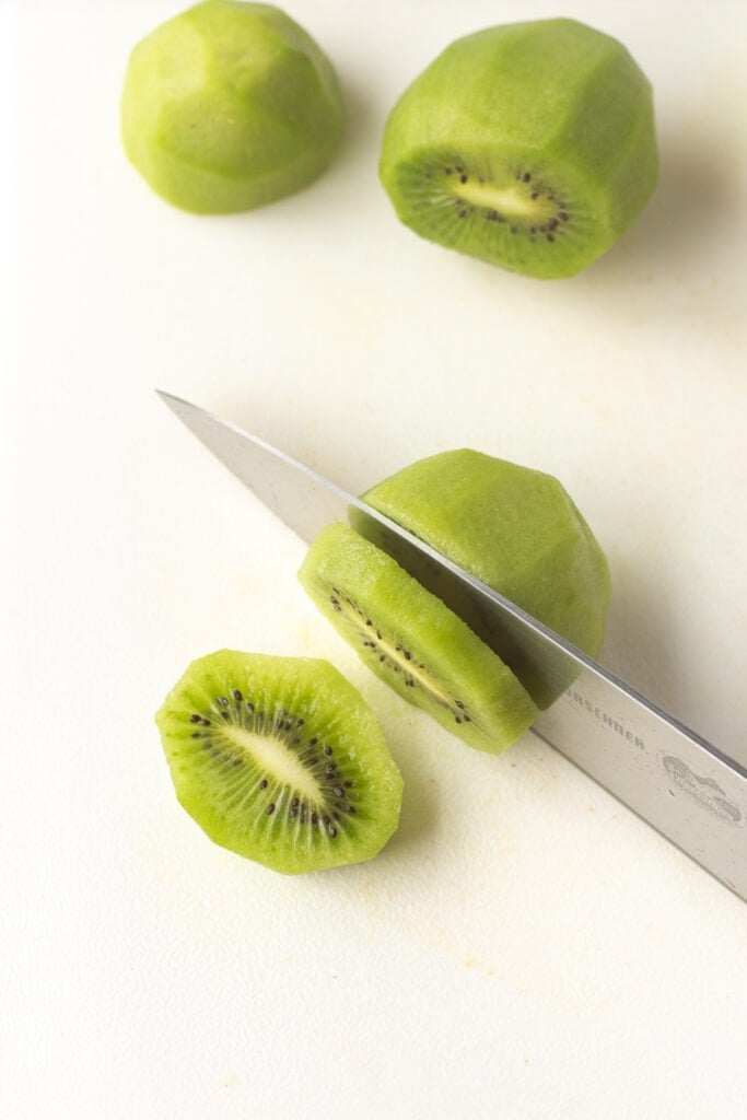 Slicing the kiwi into ¼ inch slices on a white cutting board with a large knife.