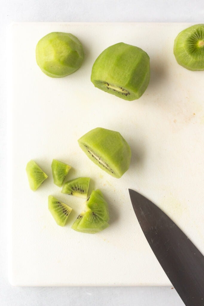 Showing a slice of kiwi that's been cut into quarters with other skinned kiwis on a large white cutting board.