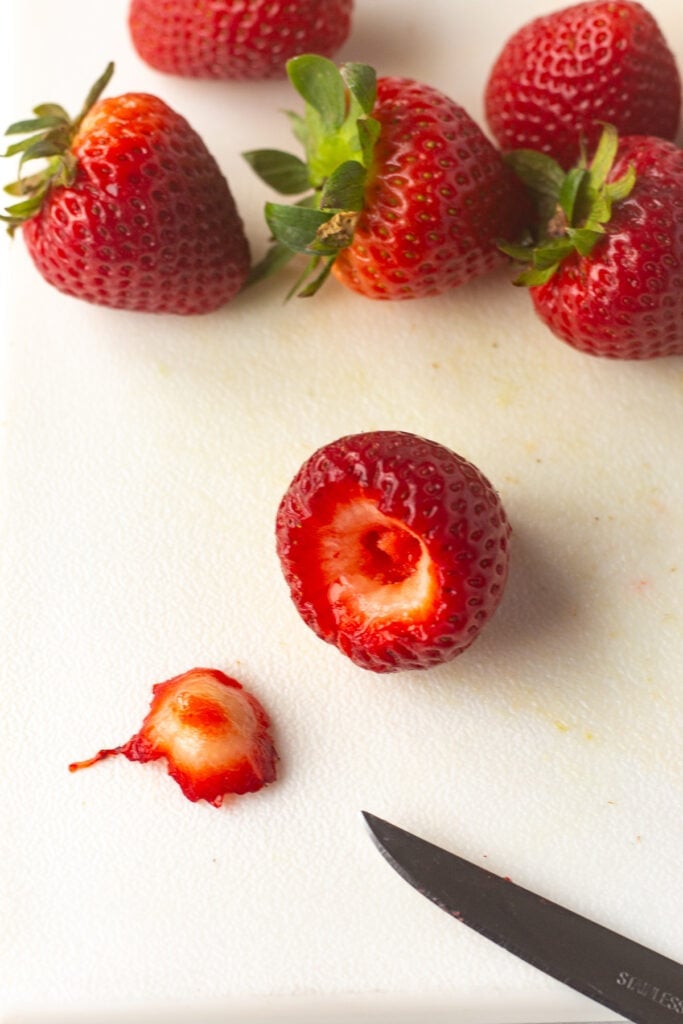 Fresh strawberries on a white cutting board, with one showing the stem cut off and a small paring knife next to it.