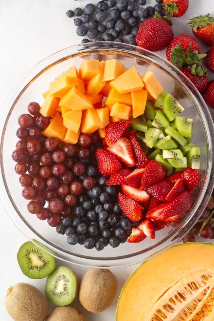 Top down shot of red grapes, blueberries, cut up cantaloupe, kiwi, and strawberries in a large clear mixing bowl next to other fresh fruit.