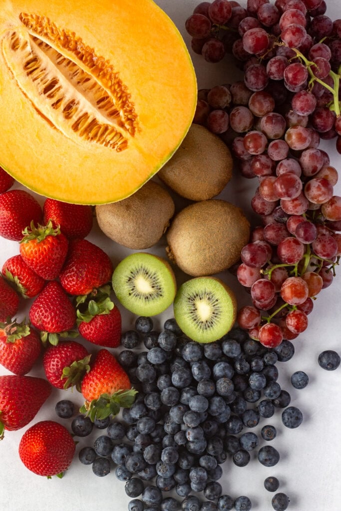 Top down shot of fresh strawberries, blueberries, kiwi, red grapes, and half a cut cantaloupe on a white background.