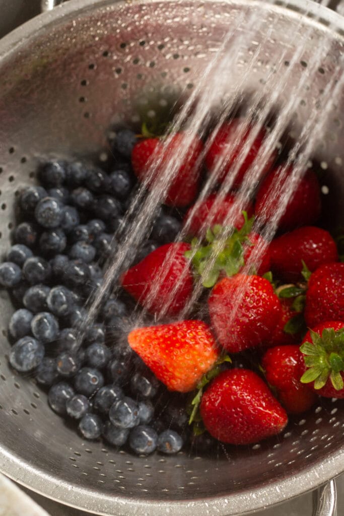 rinsing off blueberries and strawberries in a large silver colander in a sink.