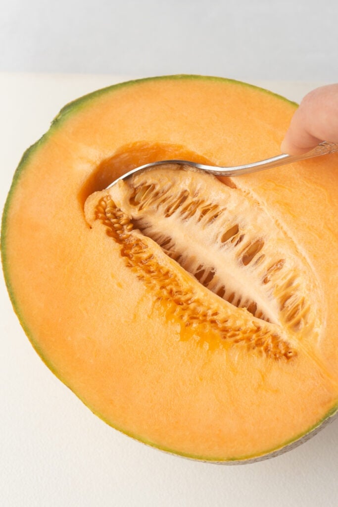A spoon scooping the seeds out of half a cantaloupe.