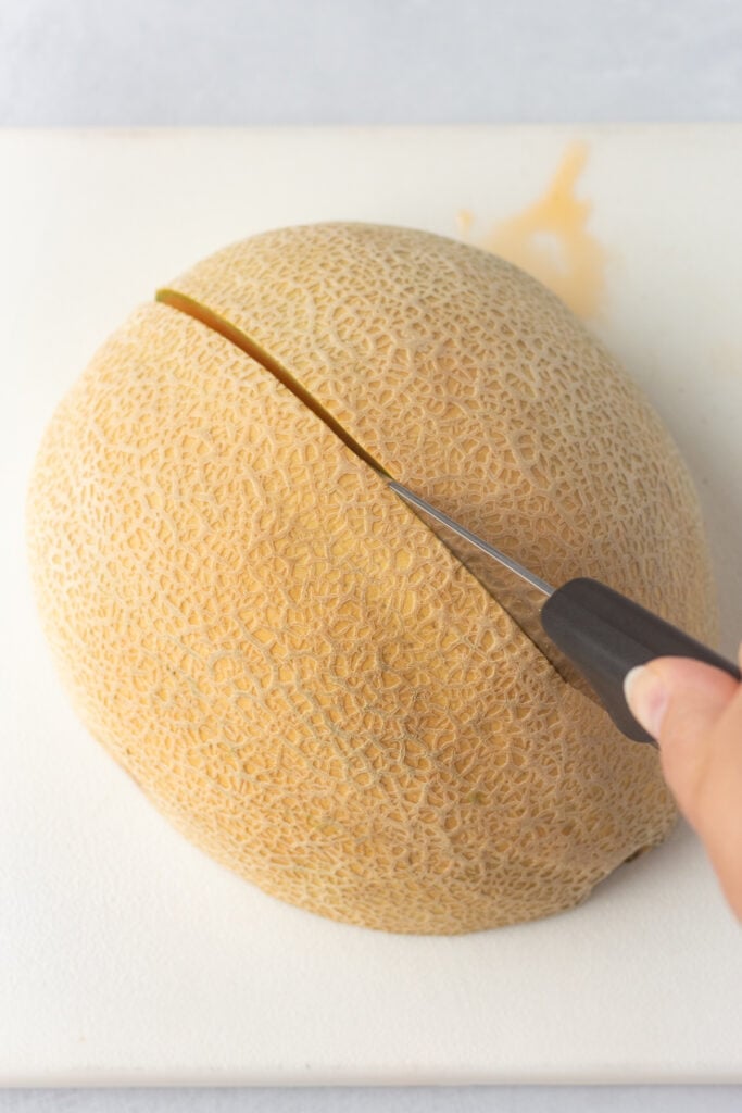 Cutting half of a cantaloupe into half again on a white cutting board.