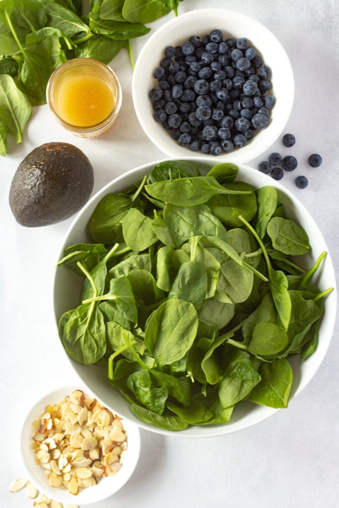 Top down shot of baby spinach, blueberries, sliced almonds, all in white bowls, next to a mason jar of dressing and a whole avocado, all on a white background.