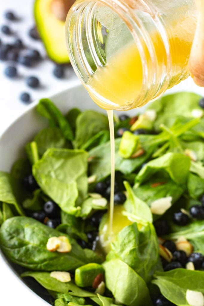 Close up of pouring white balsamic vinaigrette onto a blueberry spinach salad that's in a white serving bowl.