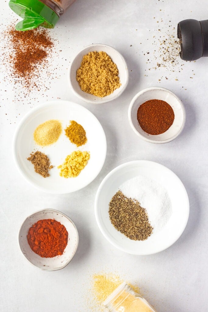 Top down shot of small bowls with different spices in them, on a white background.