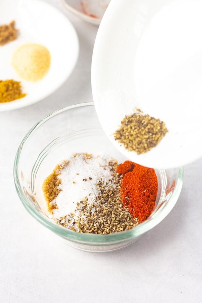 Pouring kosher salt and ground black pepper from a small white plate into a clear small bowl with other spices in it.
