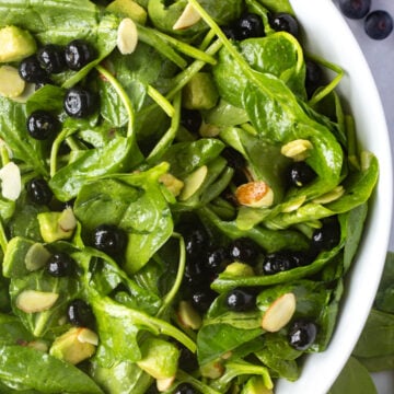 Close up of a large white salad bowl with spinach blueberry salad in it, with ingredients off to the sides.