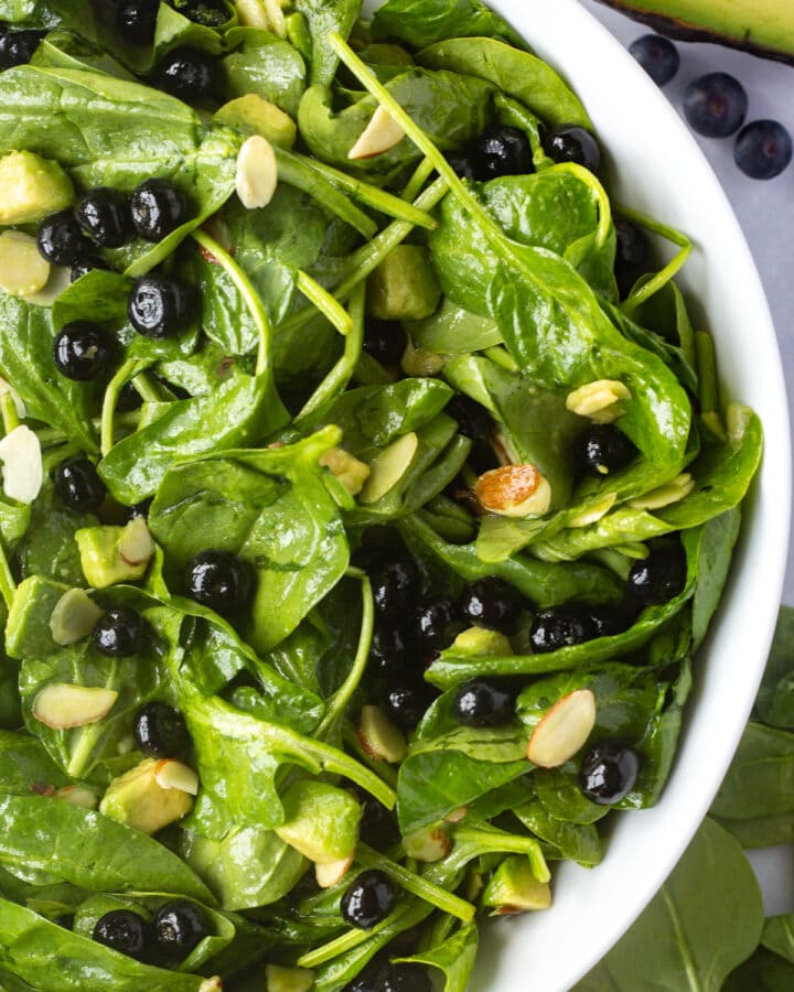 Close up of a large white salad bowl with spinach blueberry salad in it, with ingredients off to the sides.