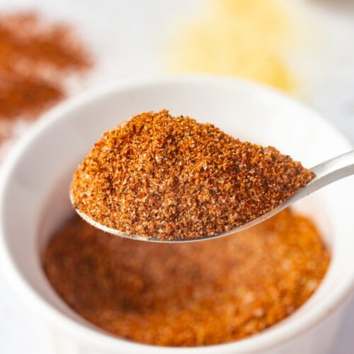Close up of a small matte silver spoon holding a large amount of brown spice rub on it, with a small white bowl of the spice rub in the background
