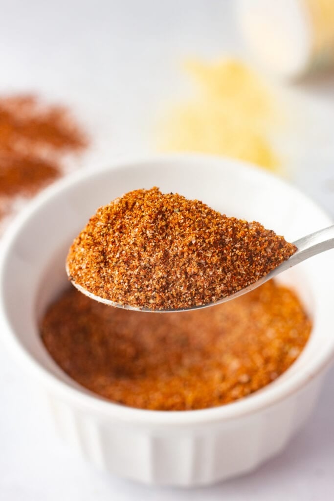 Close up of a small matte silver spoon holding a large amount of brown spice rub on it, with a small white bowl of the spice rub in the background