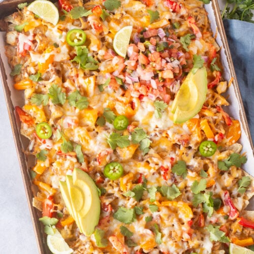 Top down shot of a sheet pan with chicken fajitas nachos on it, surrpounded by other ingredients and a blue napkin in on the right side.