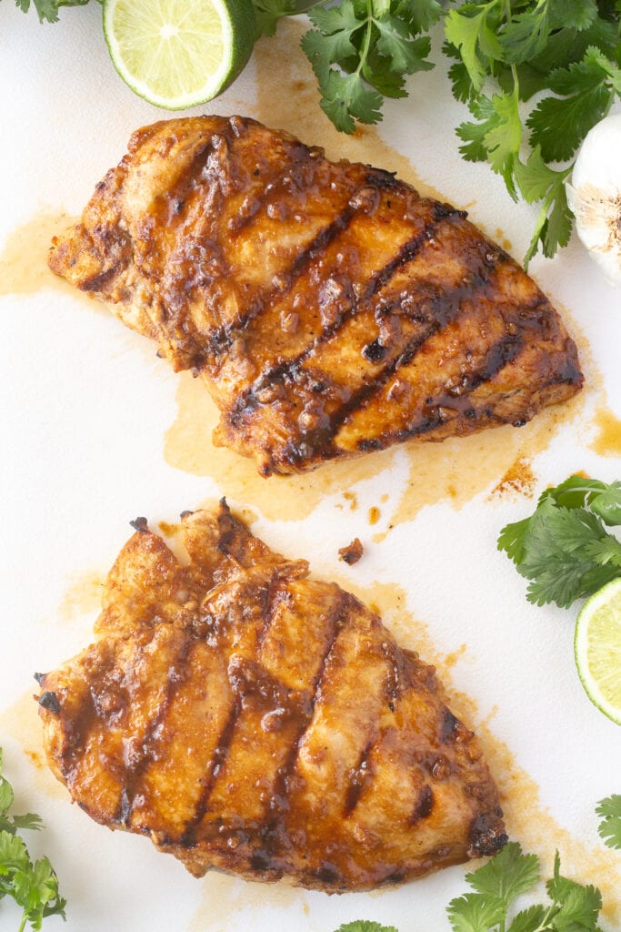 Top down shot of two grilled chicken breasts on a white cutting board with cilantro and limes surrounding them.