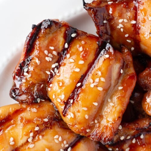 Close up of grilled boneless skinless chicken thighs with a soy honey glaze and white sesame seeds on a white platter.