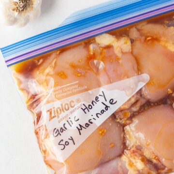 Top down shot of a bag raw boneless skinless chicken thighs in a clear ziploc bag with a brown soy marinade. A head of garlic and a honey stick with honey leaking out is in the top of the frame.