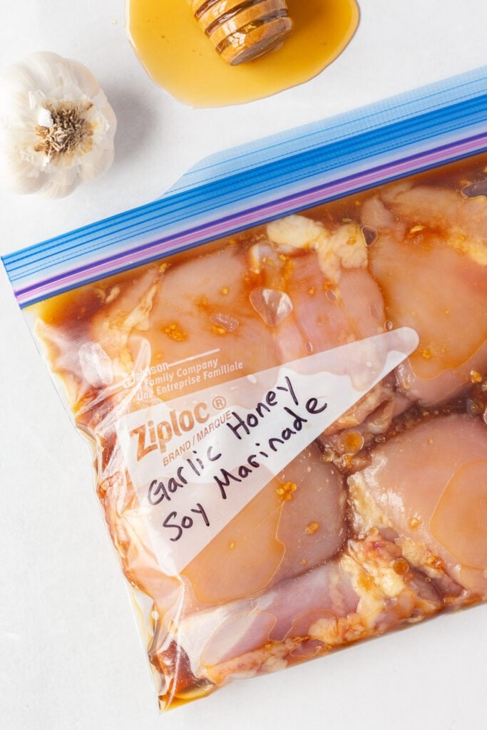 Top down shot of a bag raw boneless skinless chicken thighs in a clear ziploc bag with a brown soy marinade. A head of garlic and a honey stick with honey leaking out is in the top of the frame.