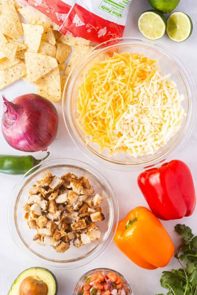 Top down shot of ingredients for chicken fajita nachos, including a bowl of shredded cheese, bell peppers, a red onion, a bowl of cooked chicken, limes, avocado, salsa, and a jalapeno.