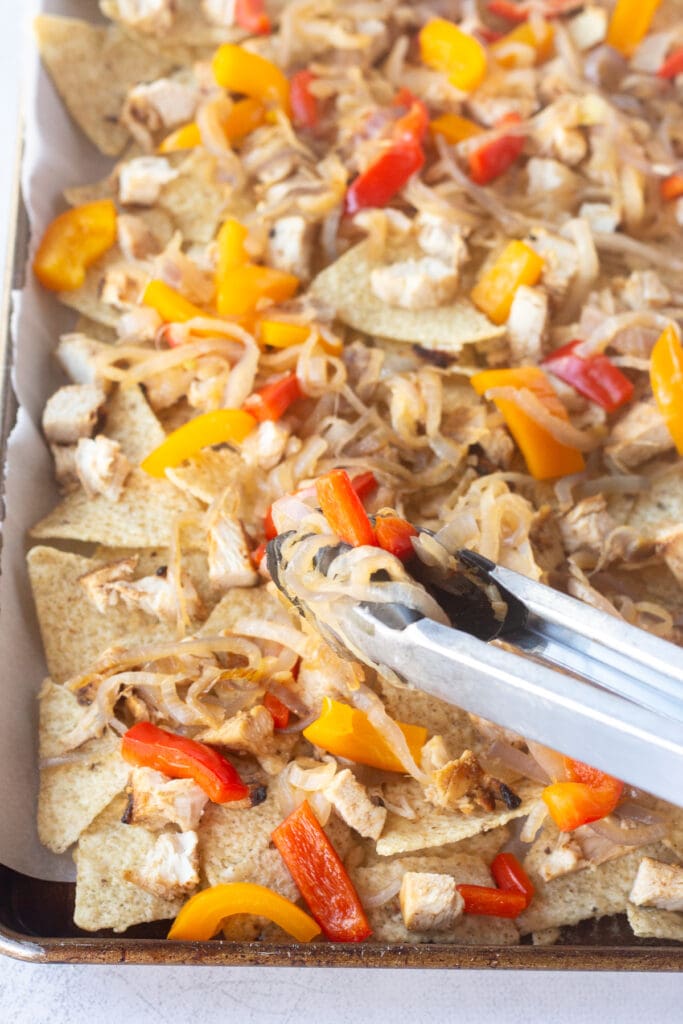 A pair of tongs placing cooked onions and bell peppers on top of a layer of tortilla chips in a sheet pan.