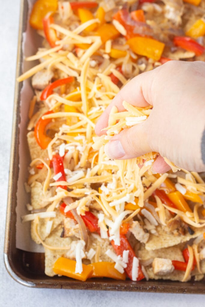 A hand placing shredded cheese on top of tortilla chips, cooked bell peppers and onions, and cooked chicken in a sheet pan.